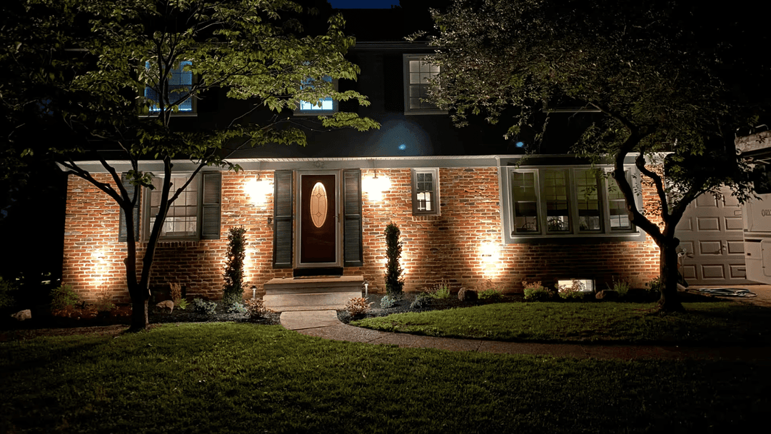 Illuminated brick house at night with landscaped yard and tree in front.