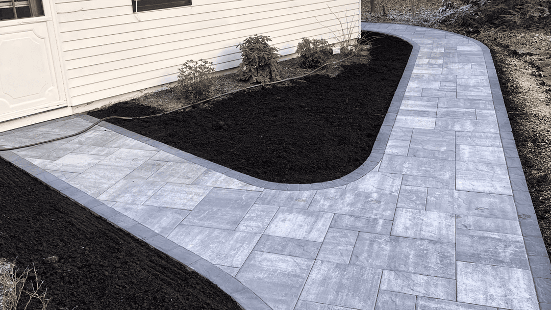 Curved gray stone pathway leading to a house, bordered by dark soil and shrubs.