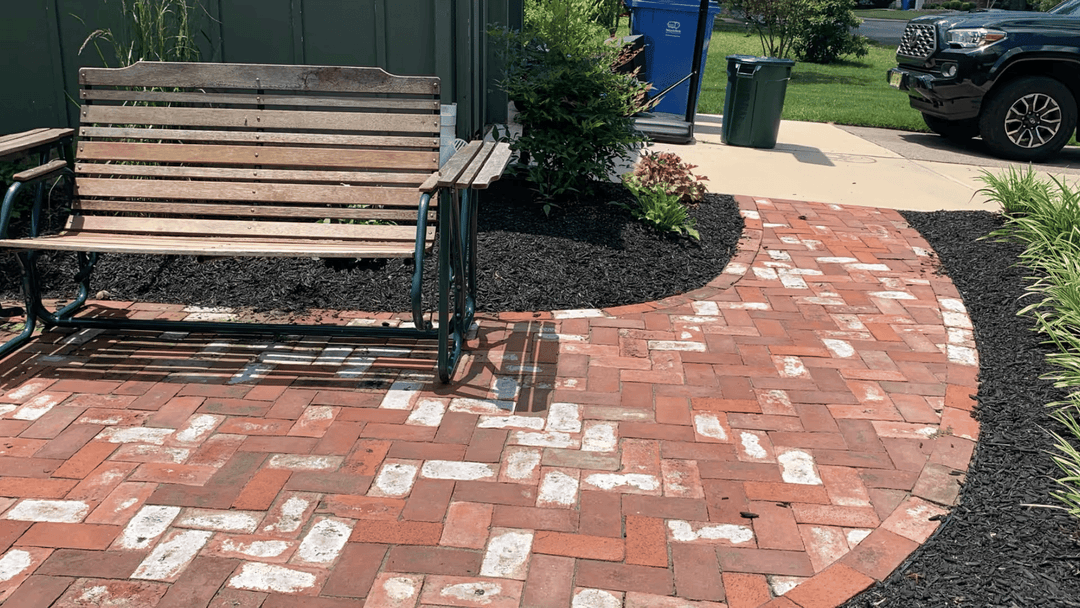 Wooden bench on a brick pathway with landscaping and a parked vehicle in the background.
