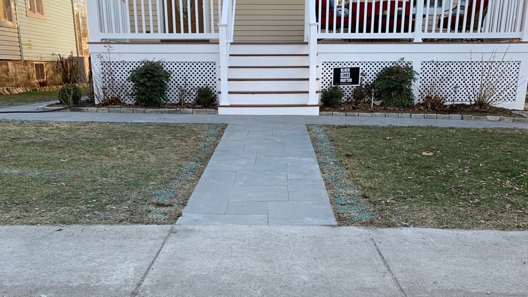 Stone walkway leading to a house with porch steps, bordered by grass and shrubs.