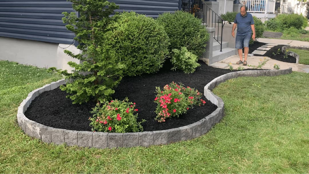 Man tending to a landscaped garden with flowers and mulch near a home.