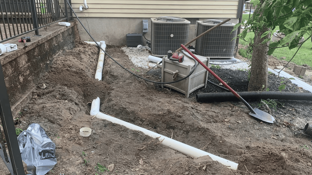 Excavated area showing PVC pipes, tools, and air conditioning units in a residential yard.