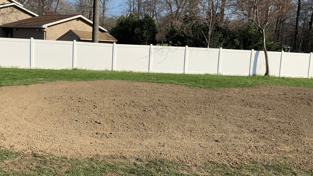 Cleared dirt patch in lawn with a white fence and house in background, ready for landscaping.