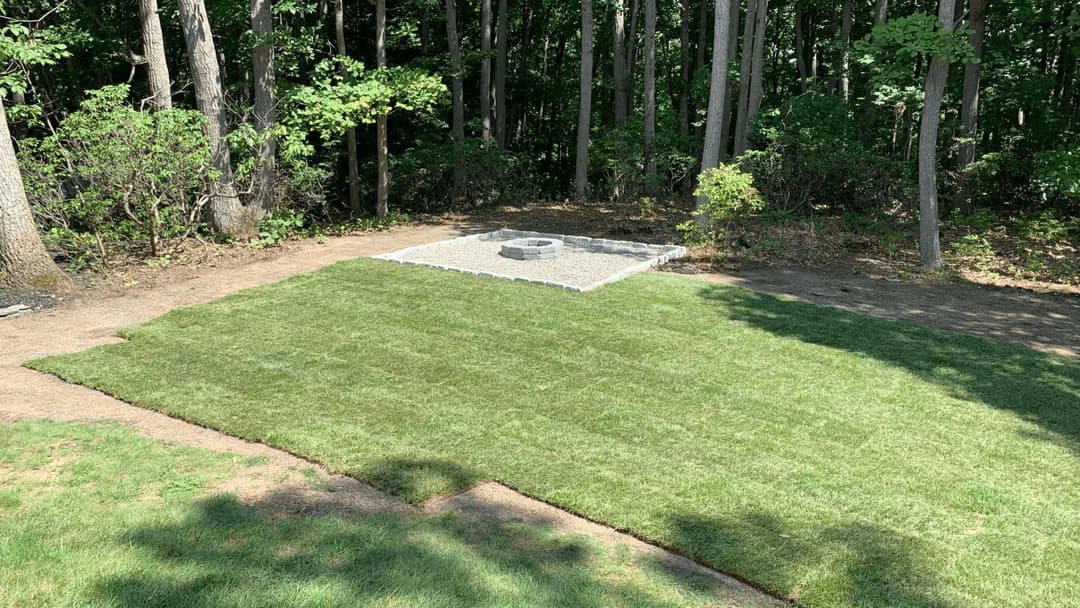 Lush green lawn with a stone fire pit area surrounded by trees in a wooded setting.