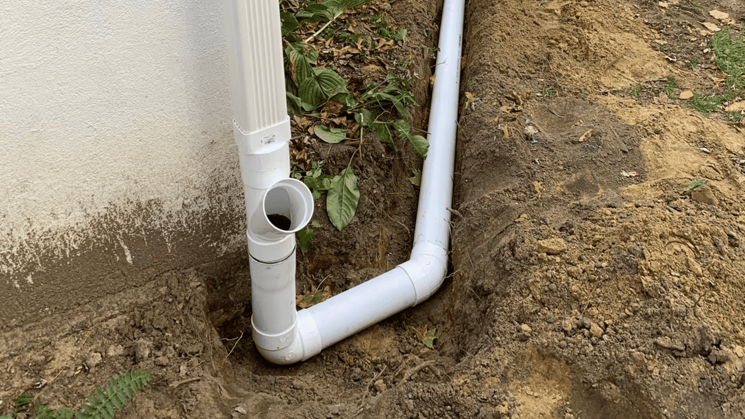 White PVC drainage pipe installed in soil, directing water away from a building foundation.