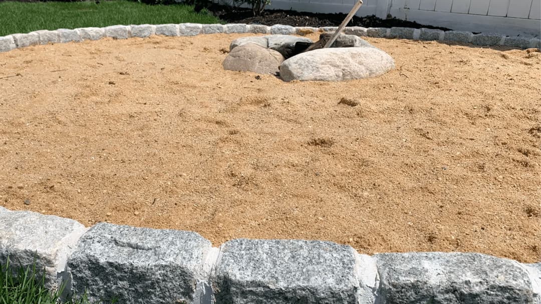 Sand pit with stone border and a rock centerpiece in a backyard setting.