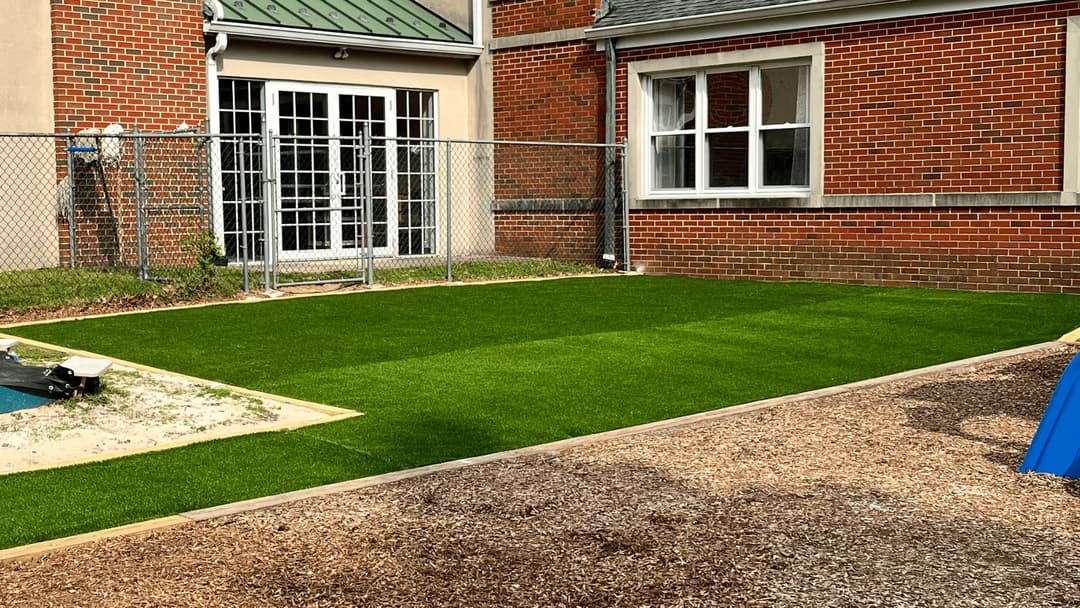 Artificial turf lawn adjacent to a brick building with a fenced area and playground surface.