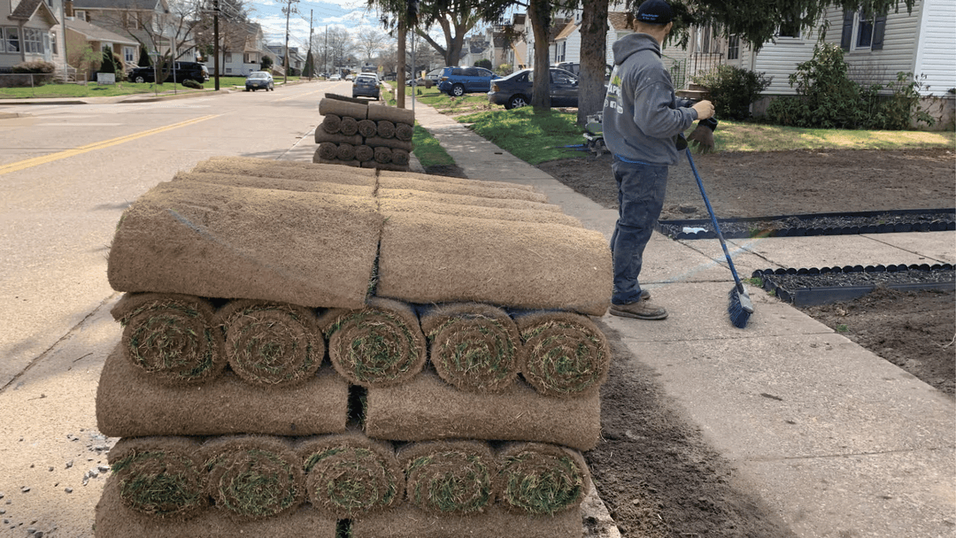 Person rolling out fresh sod along a residential street, stacked rolls visible nearby.