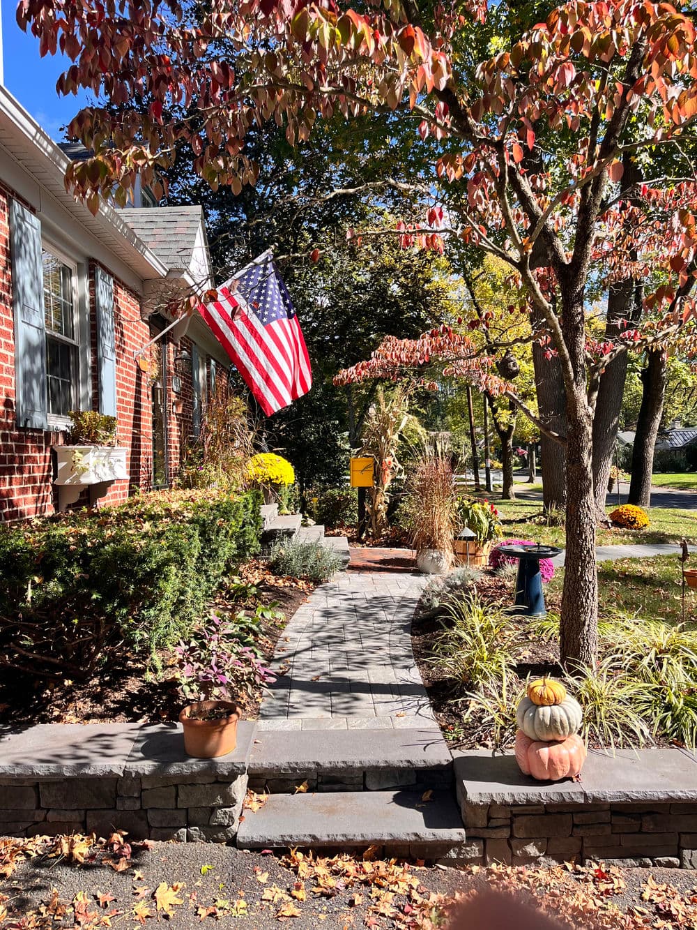 American flag next to a brick house in autumn, with pumpkins and colorful foliage along the pathway.