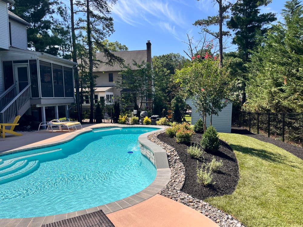 Backyard pool with landscaped garden, trees, and a cozy sitting area on a sunny day.