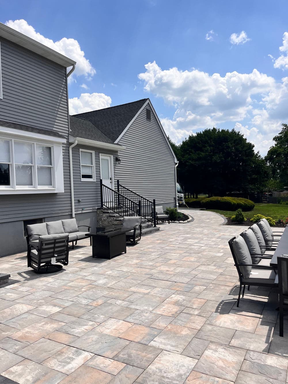 Spacious backyard patio with seating, dining area, and lush greenery under a blue sky.