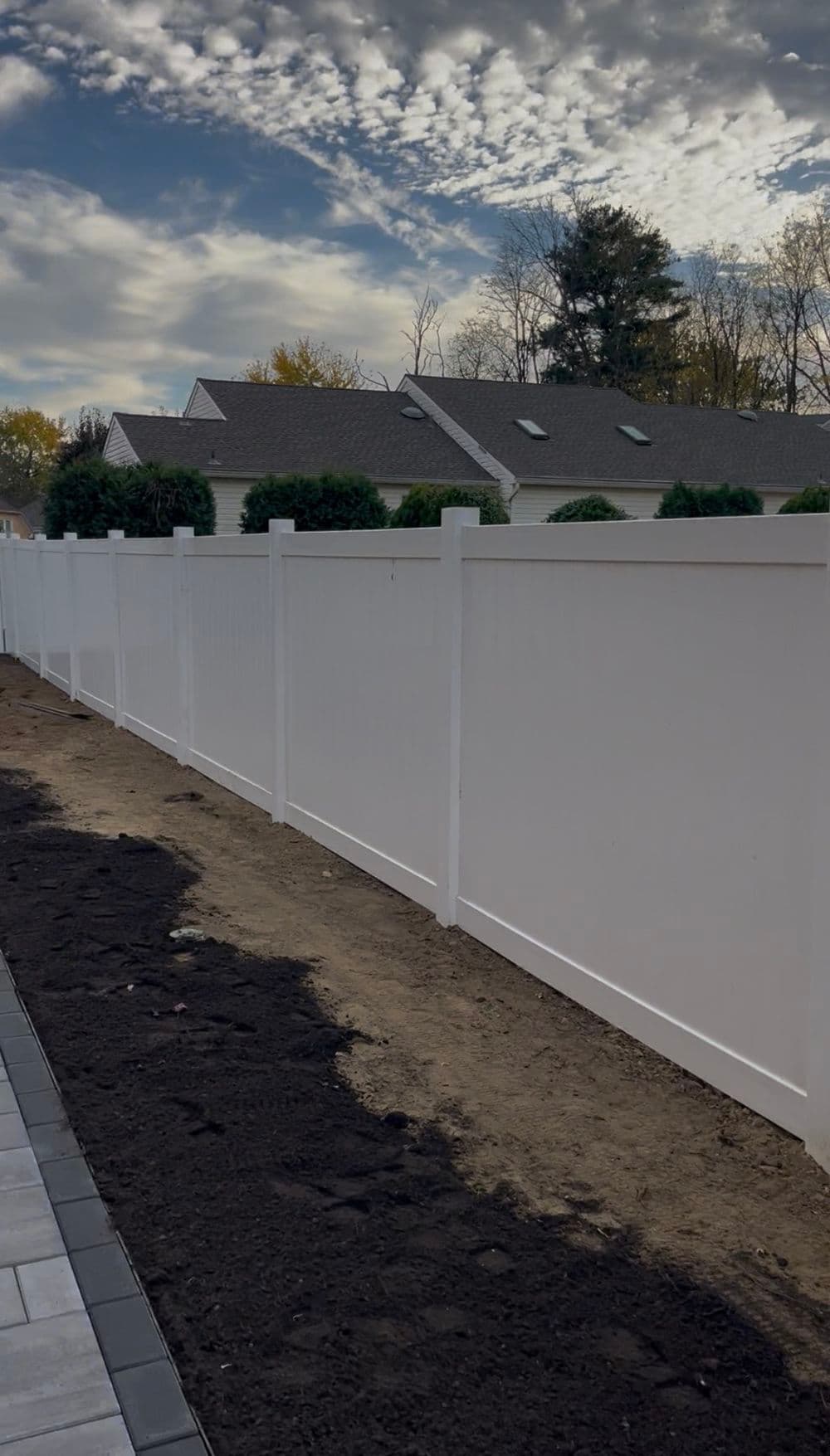 White vinyl fence along a landscaped yard under a cloudy sky, featuring a freshly dug area.